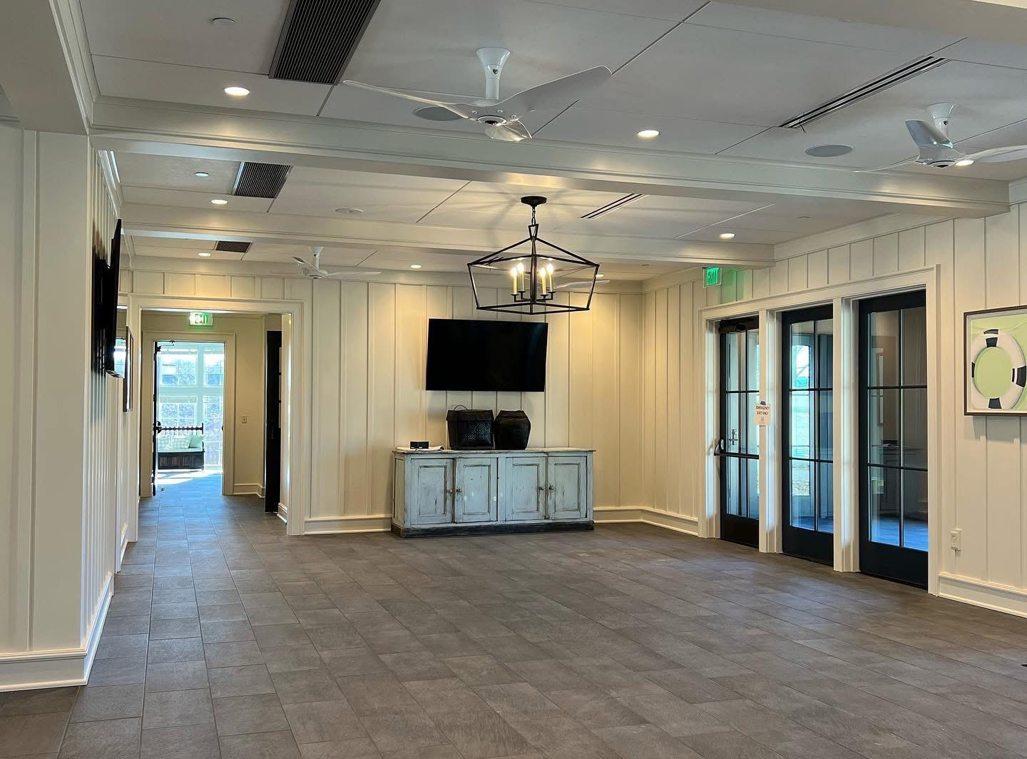 A spacious, empty room with light-colored walls, gray tile flooring, double glass doors, a wall-mounted TV above a cabinet, large ceiling lights, and ceiling fans; sunlight enters through a hallway on the left—perfect for an Asheville painter’s studio.