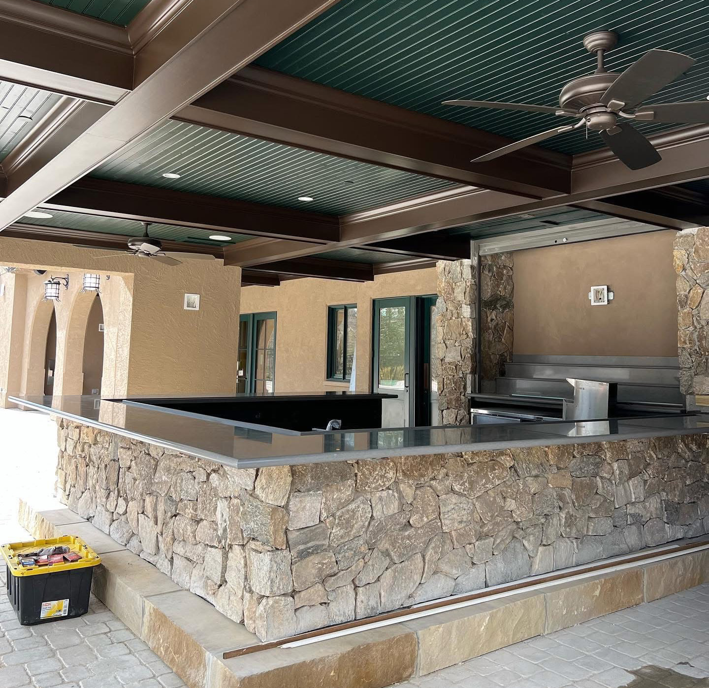 Spacious outdoor kitchen with a stone bar, stainless steel grill, and green ceiling with fans—perfect for Asheville gatherings. Tan stucco walls surround the space, while a yellow and black toolbox hints at recent painter work in the foreground.