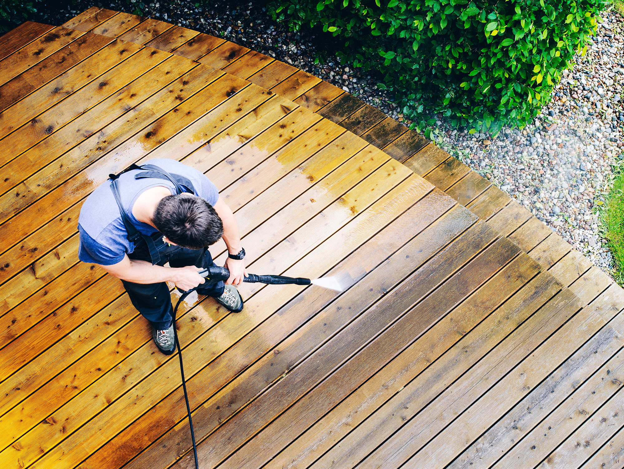 A person in a blue shirt and overalls uses a pressure washer to clean a wooden deck in Asheville; green bushes, gravel, and fencing are visible along the deck’s edge.