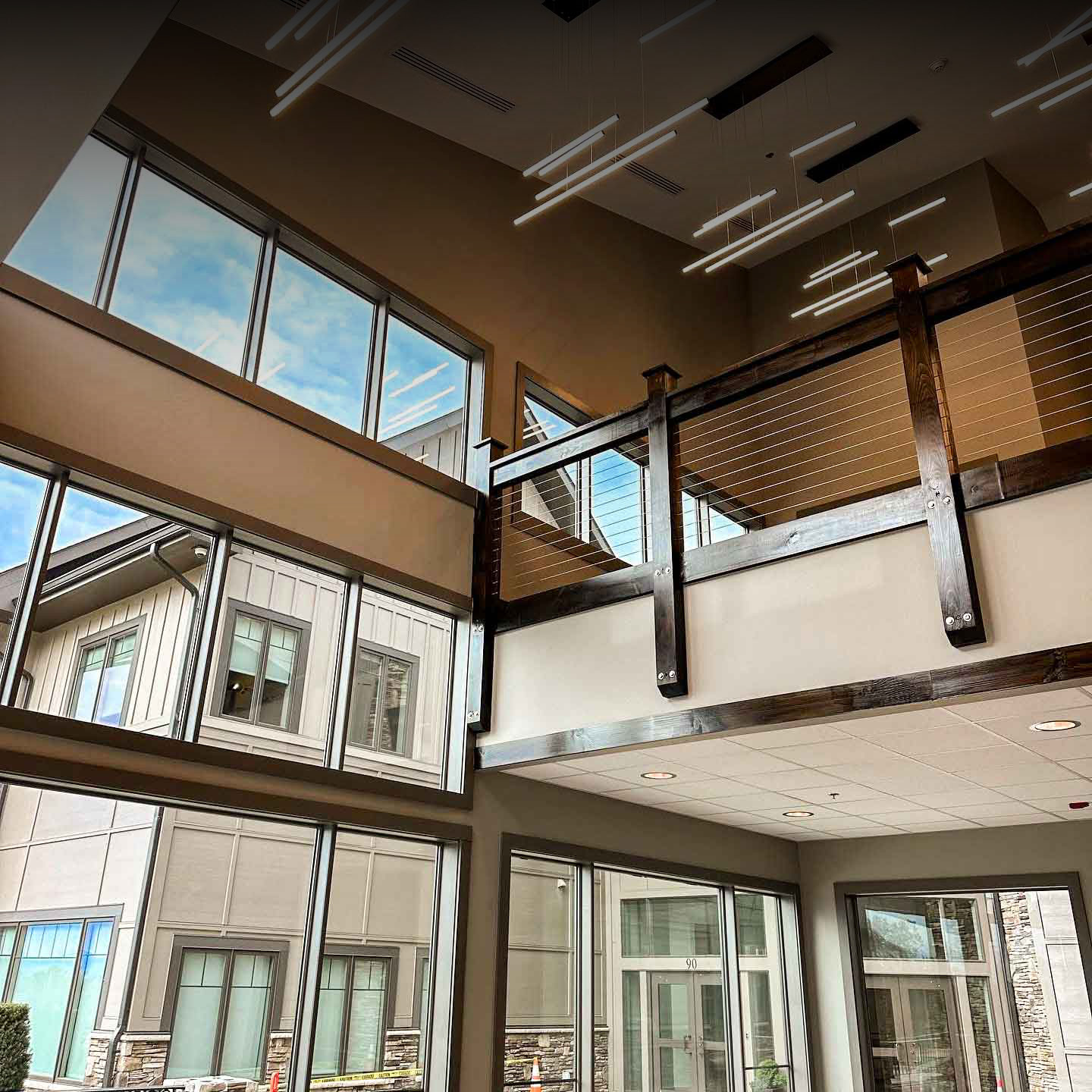 Modern building interior with large windows, natural light, wood and cable railing on an upper level, and fluorescent ceiling lights. Exterior of nearby building visible through windows.