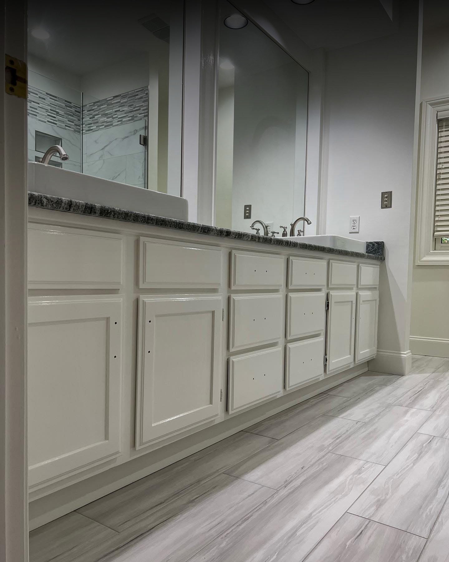 White bathroom cabinets with no door handles, beneath a granite countertop with two sinks and mirrors. The floor has grayish wood-look tiles, and a shower is visible in the background. Walls and lighting give a modern feel.
