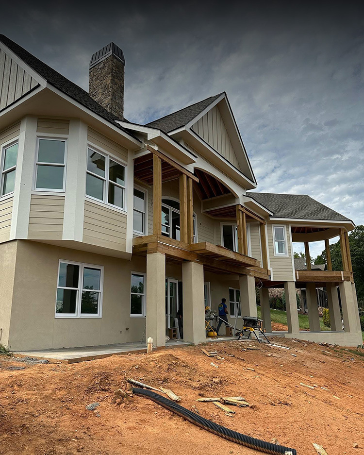 Two-story house under construction with beige siding, large windows, stone chimney, and wooden posts on the porch. Dirt and construction materials are scattered in the yard. Two people work near the entrance.