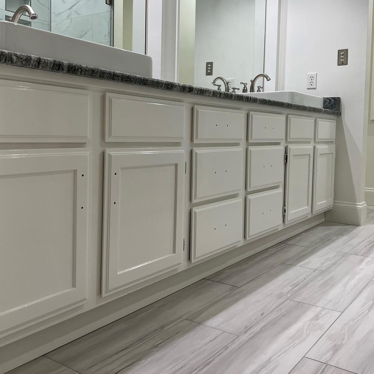 White bathroom cabinets without handles are shown beneath a dark granite countertop with two sinks. The clean, modern space features painter-fresh white walls, light gray marble-like tiles, and stylish touches inspired by Asheville design.