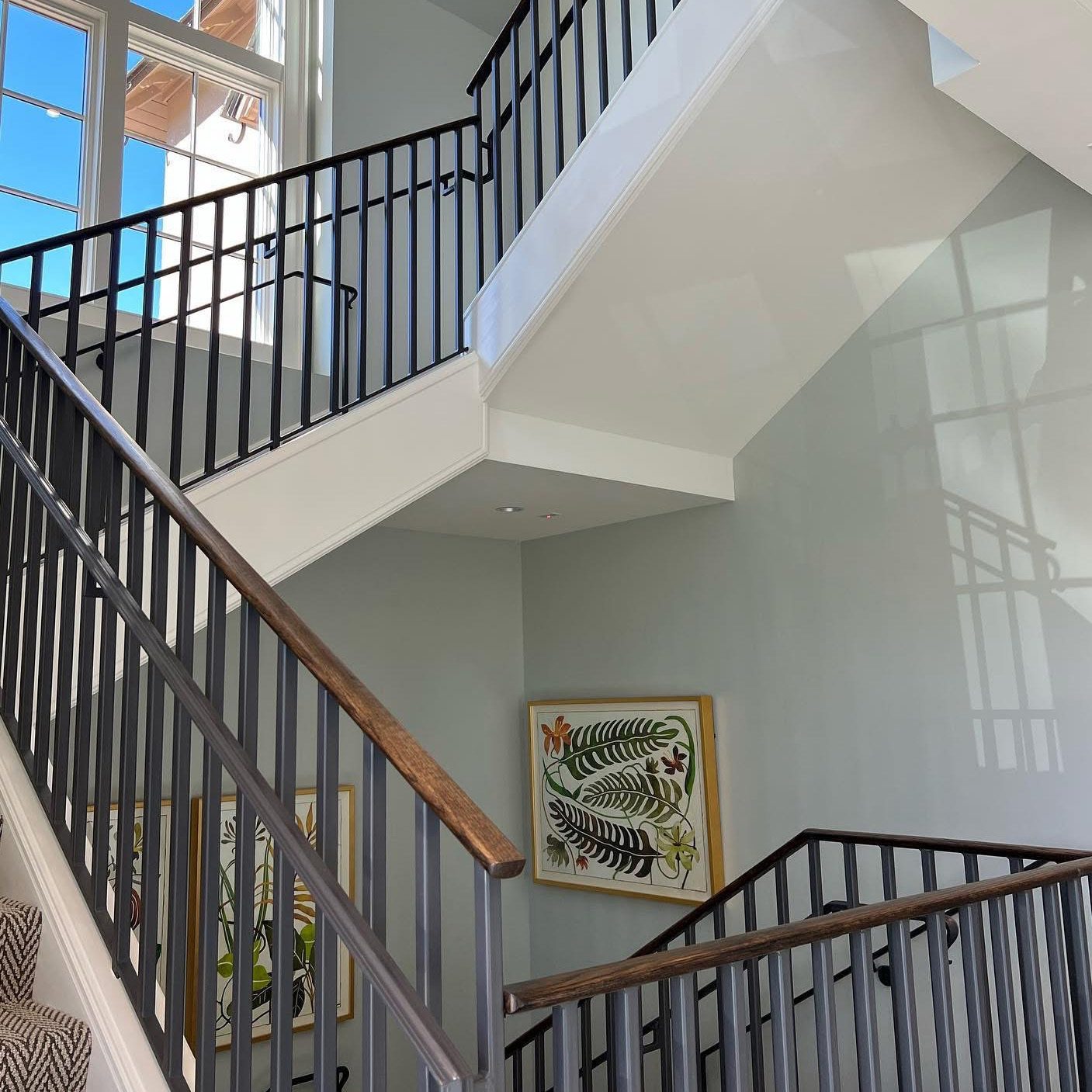 A modern indoor staircase with black metal railings, wooden handrails, and light-colored walls. Large windows let in natural light. Framed botanical artwork by a local Asheville painter hangs on the walls by the stairs.