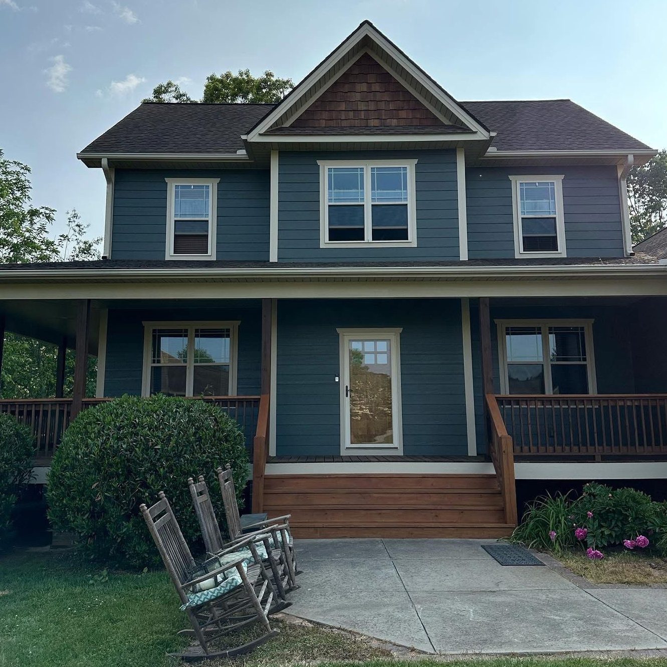 A two-story blue house with a covered porch and wooden railings, surrounded by green bushes. Four rocking chairs are arranged on the lawn near the front steps, perfect for relaxing after a day’s work by the painter finishing up inside.
